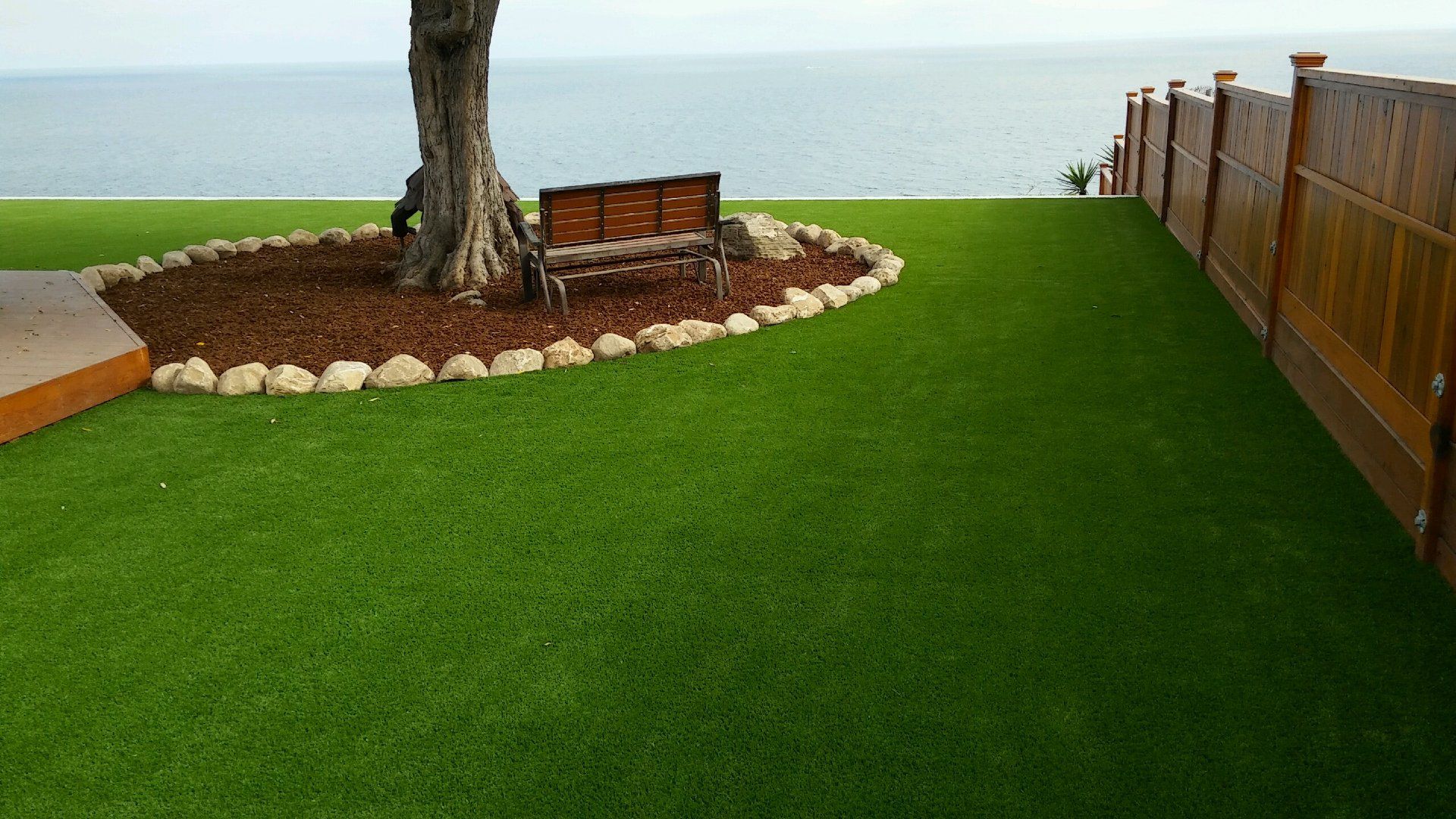 Backyard lawn with a tree, stone border, bench, and ocean view beyond a wooden fence