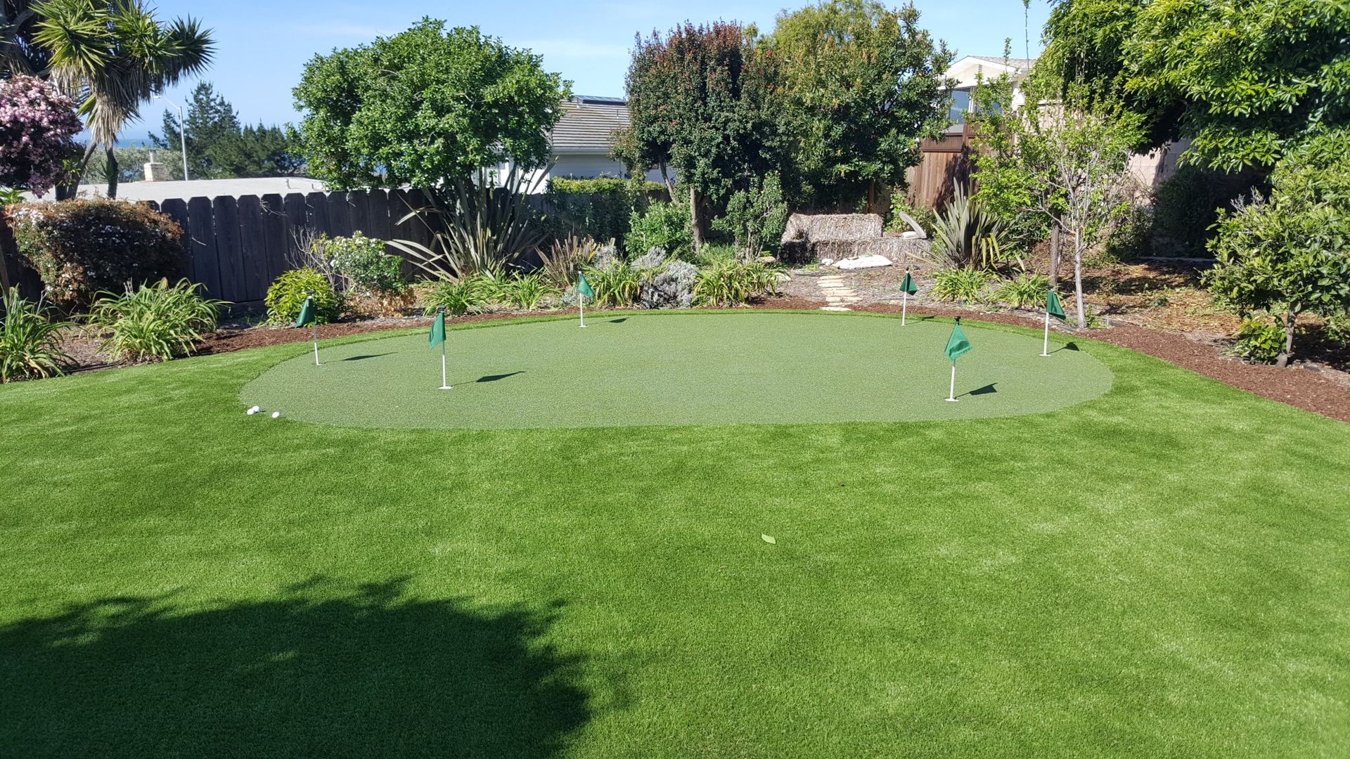 A green lawn with a circular putting green, surrounded by shrubs, trees, and a garden fence.
