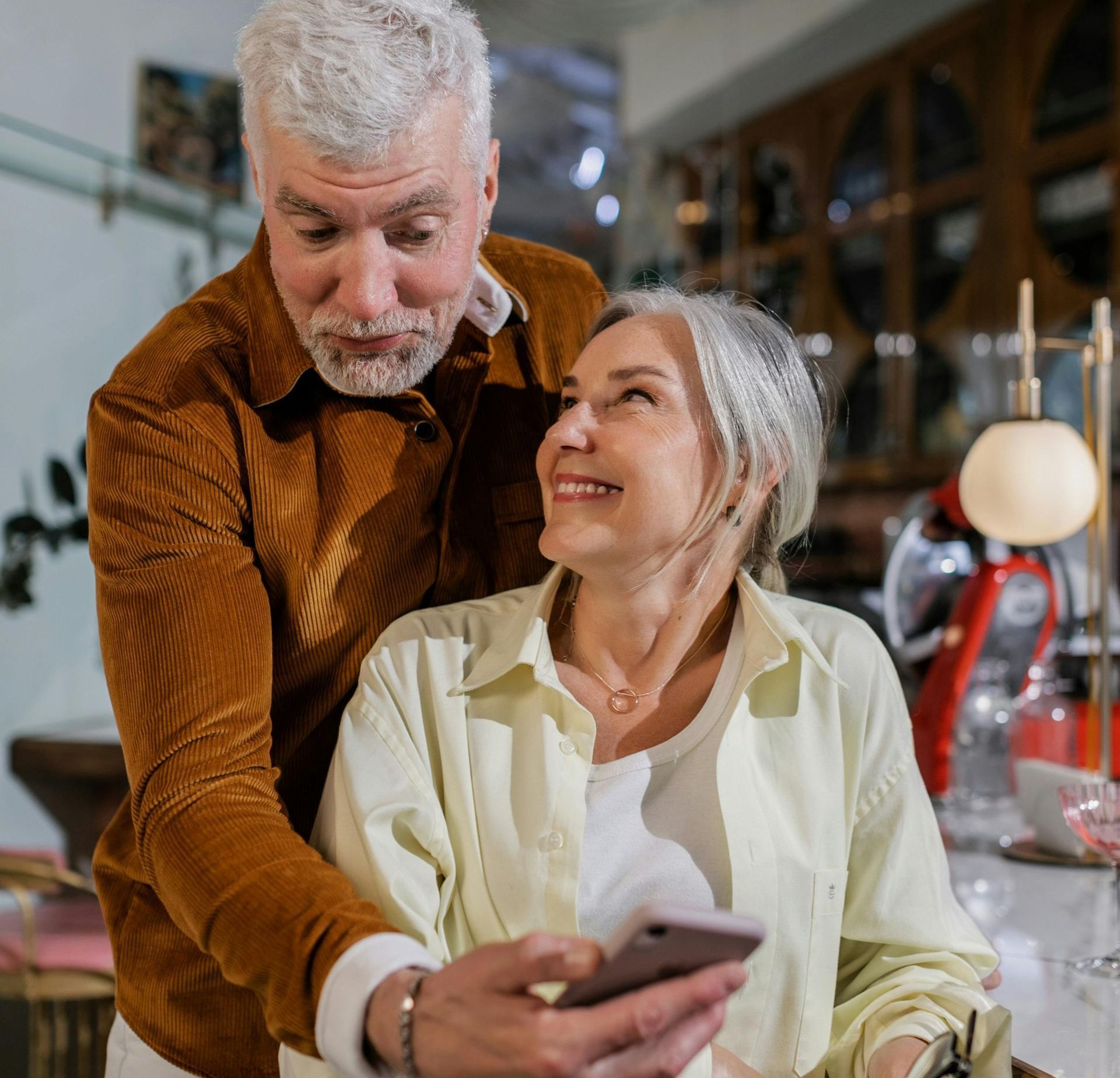 Mature couple looks at a phone together indoors. Man with brown jacket, woman with light shirt smiles.