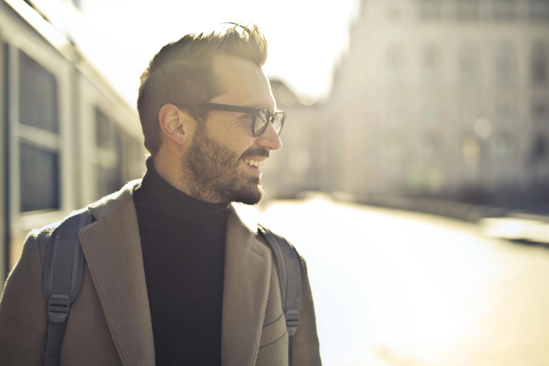 Man with glasses, a beard, and a black turtleneck smiles, wearing a tan coat, standing outdoors with a backpack.