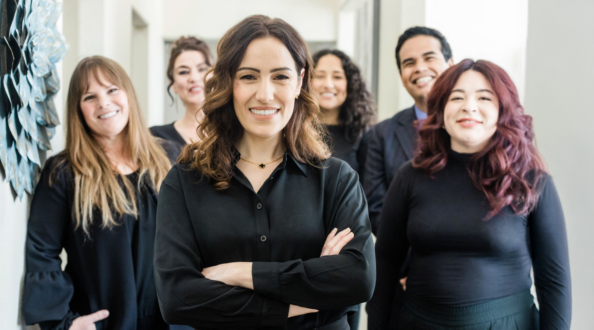 Five coworkers standing together indoors, smiling, with arms crossed in a bright office setting