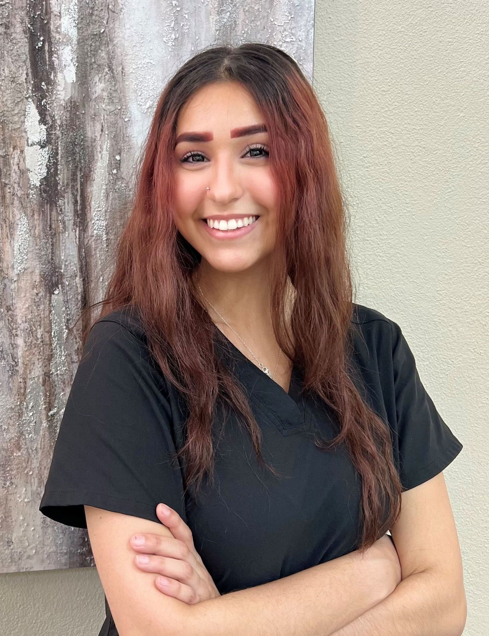 A woman in a black scrub top is smiling with her arms crossed in front of a painting.
