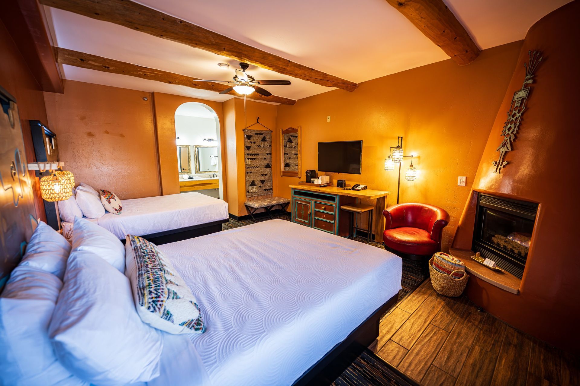 Cozy bedroom with two white beds, warm orange walls, wood beams, desk, lamp, and fireplace