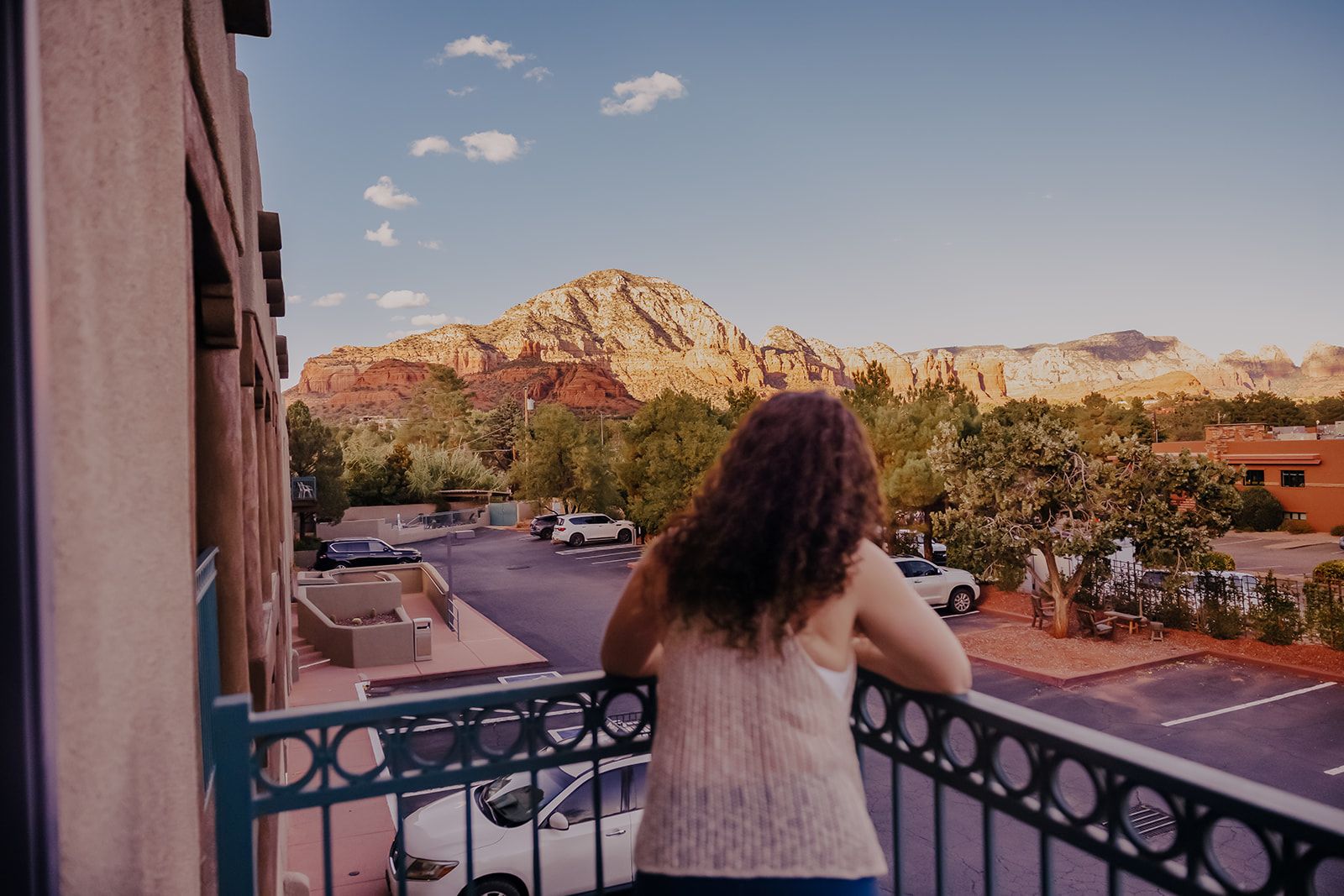Person on a balcony looking at a mountain at sunset, with a street and parked cars below.