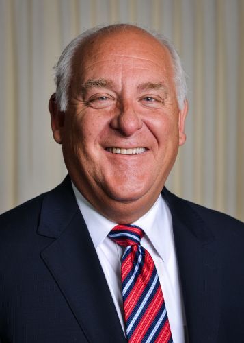 Man in navy suit, white shirt, and red/blue striped tie, smiling in front of a neutral backdrop.