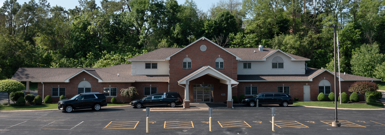 A one-story brick building with a covered entrance, several parked cars, and a surrounding parking lot and trees.