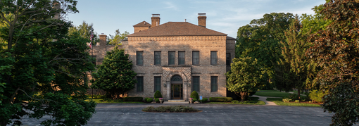 A grand stone mansion with a gray roof is surrounded by trees in the daytime.