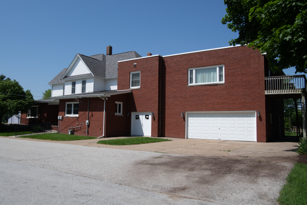 Two-story building: white farmhouse with brick addition, two-car garage, and gravel driveway on sunny day.