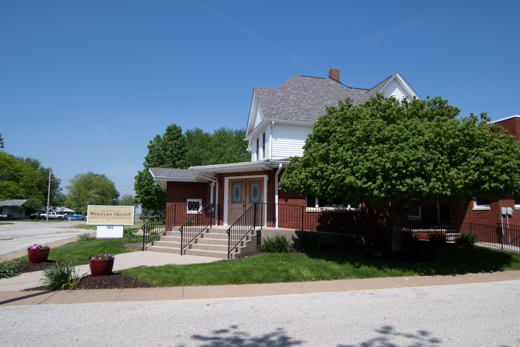 White building with a brick addition. Steps lead to the front doors, with a sign and flower pots in front.