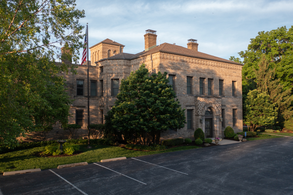 Stone building with multiple windows, chimneys, and an American flag. Trees and a parking lot are in the foreground.