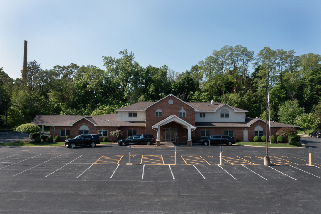 Brick building with a parking lot. Trees in the background, a tall industrial chimney. Several parked cars.