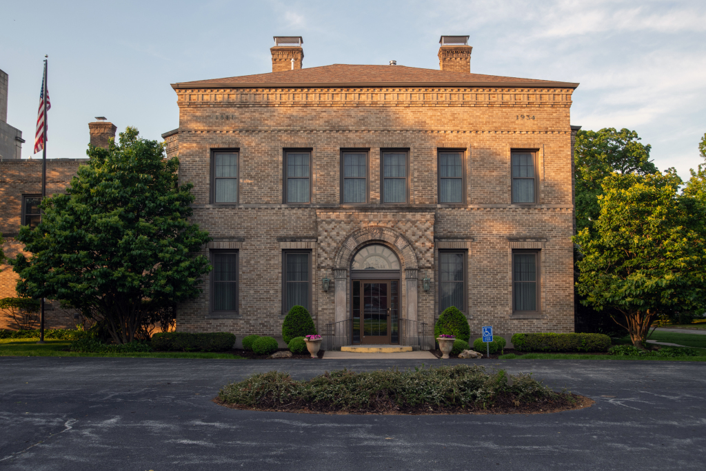 Brick building with multiple windows and central arched doorway. A flag pole and trees are in the foreground.
