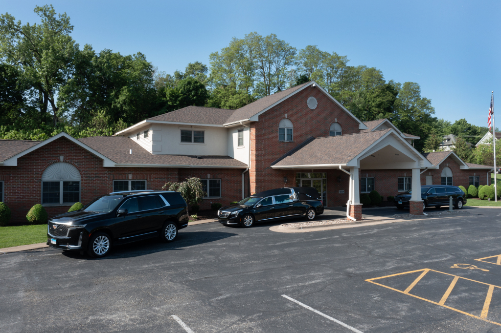 Funeral home with hearse and two limousines parked out front. Brick building with brown roof.