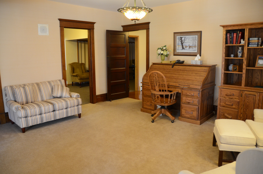 Room with tan carpet and walls, wooden desk, bookshelf, and two sofas.