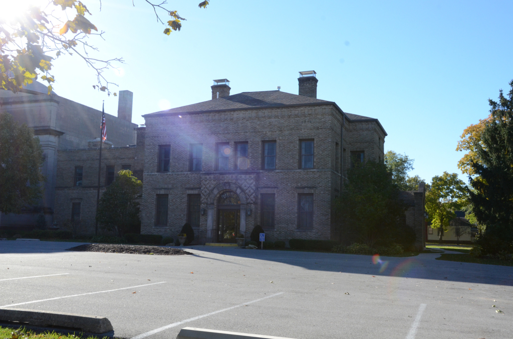 Stone building with a central arched entrance, in a parking lot on a sunny day.