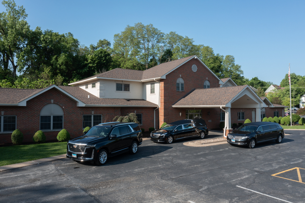 Black vehicles parked outside a brick building with a covered entrance; trees in the background, sunny day.