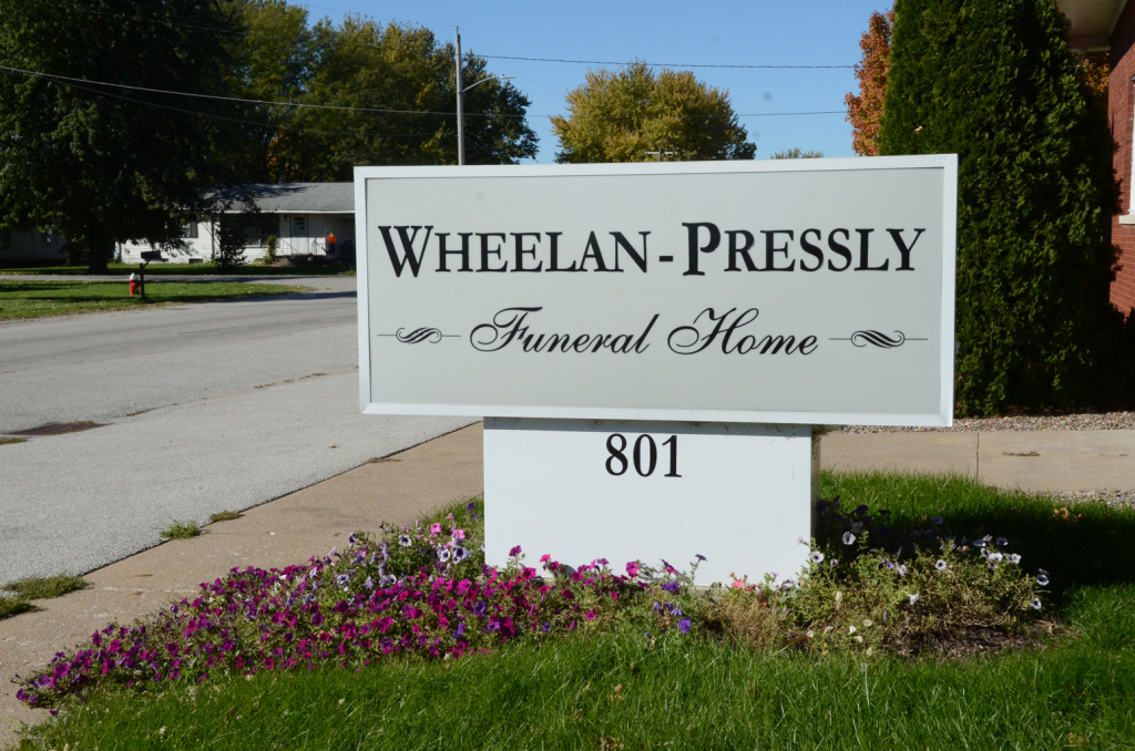 Sign for WHEELAN-PRESSLY Funeral Home at 801, with flowers in front and residential street in background.