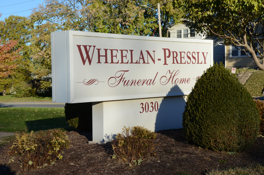 Sign for Wheelan-Pressly Funeral Home, tan and white, with address 3030; surrounded by bushes and trees.