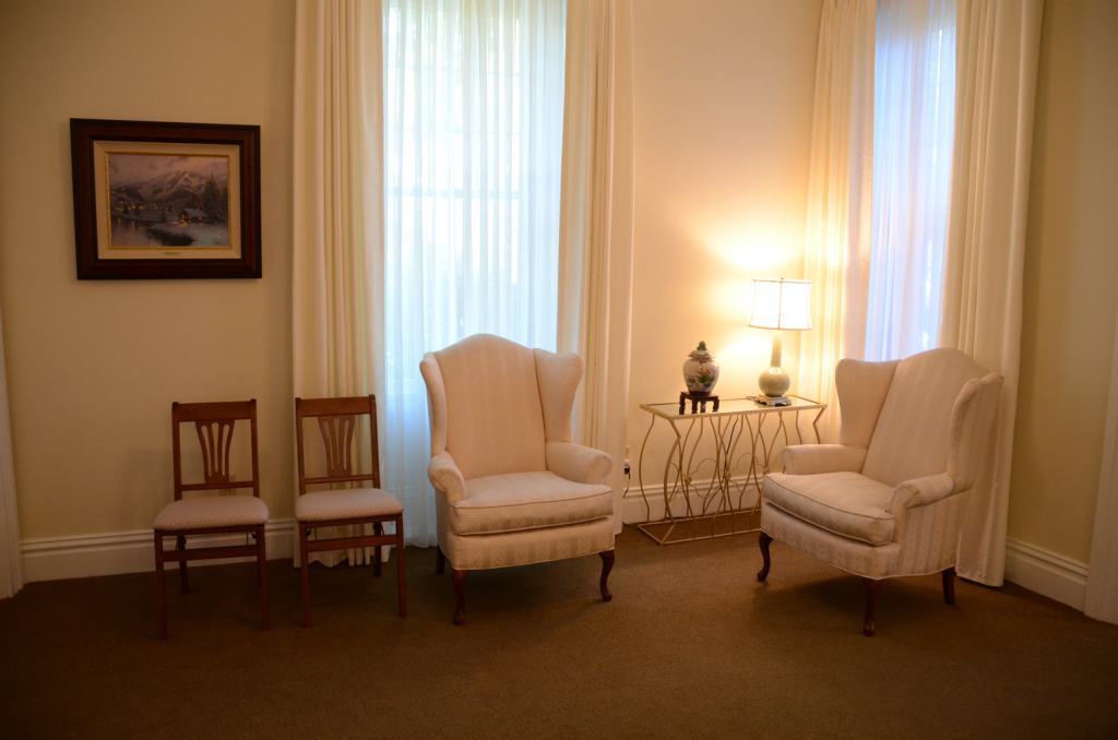 A room with two armchairs, two wooden chairs, and a small table between them, next to curtains and a painting.