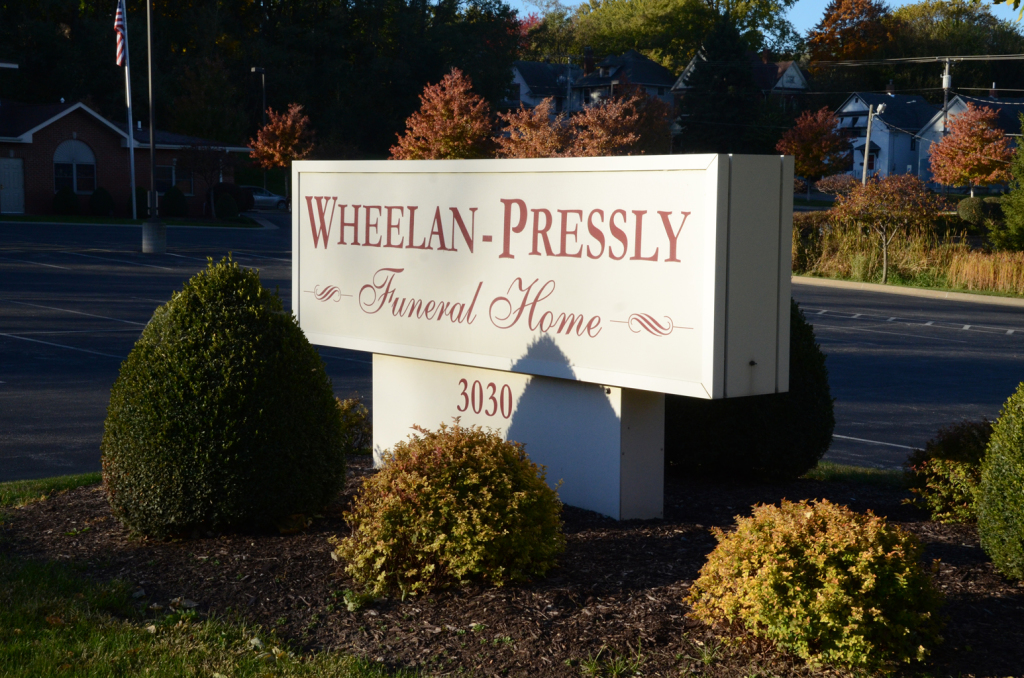 Sign for Wheelan-Pressly Funeral Home; white with brown lettering, bushes in foreground.