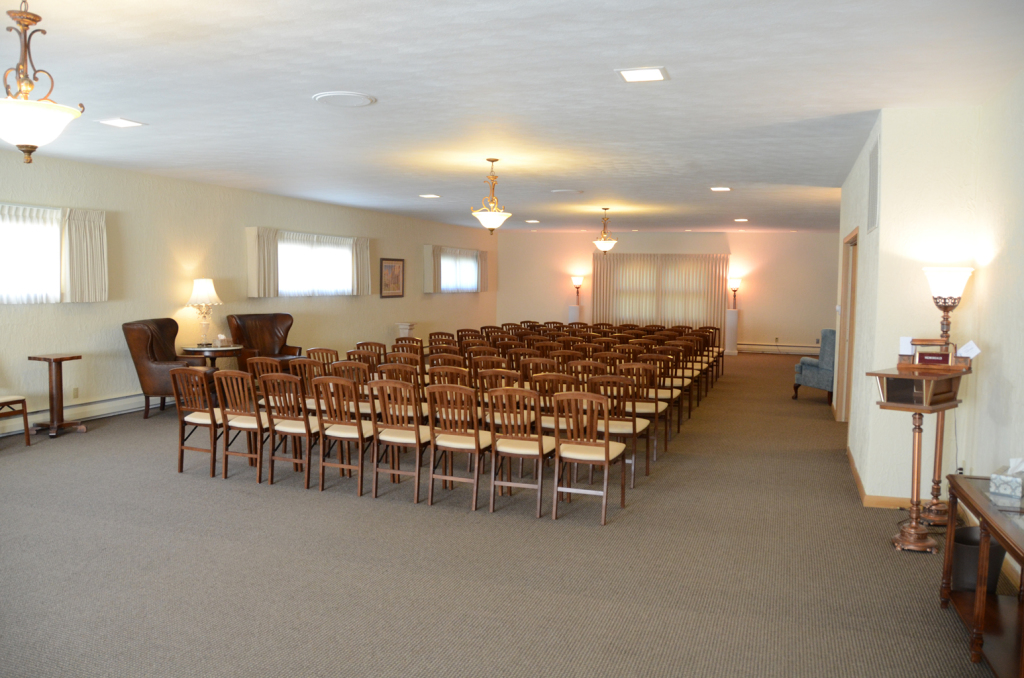 Rows of empty wooden chairs in a carpeted room; likely a funeral home.