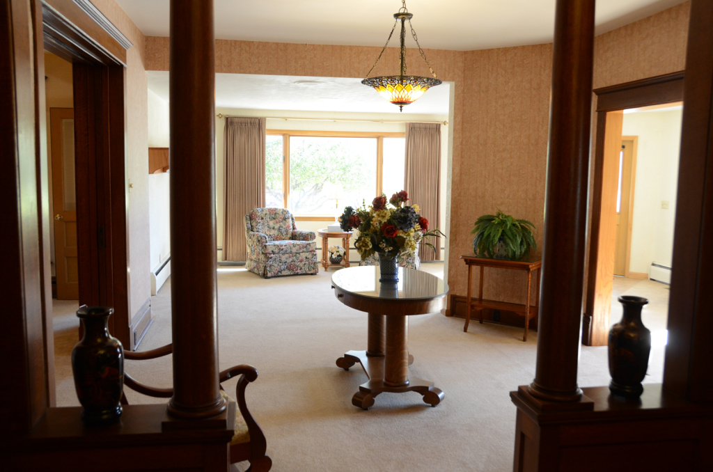 Interior view of a living room with decorative pillars, a round table with flowers, and a window with a chair.