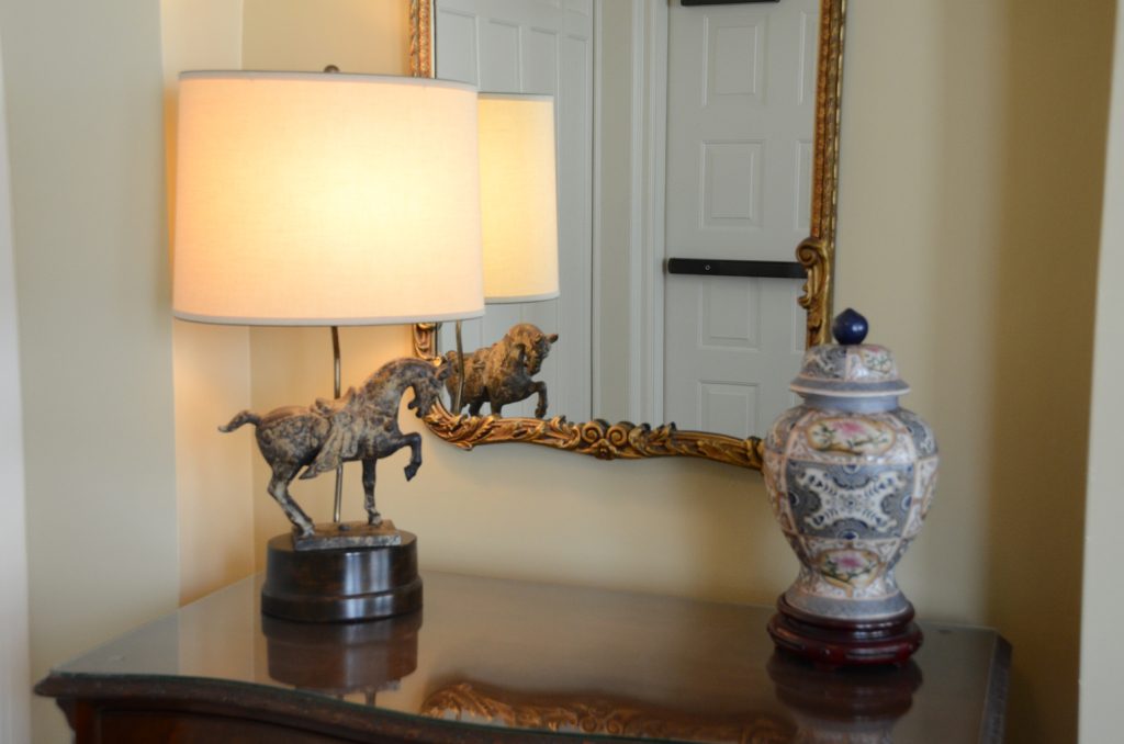 A lamp with a horse statue base, a jar, and a mirror on a dark wood table.