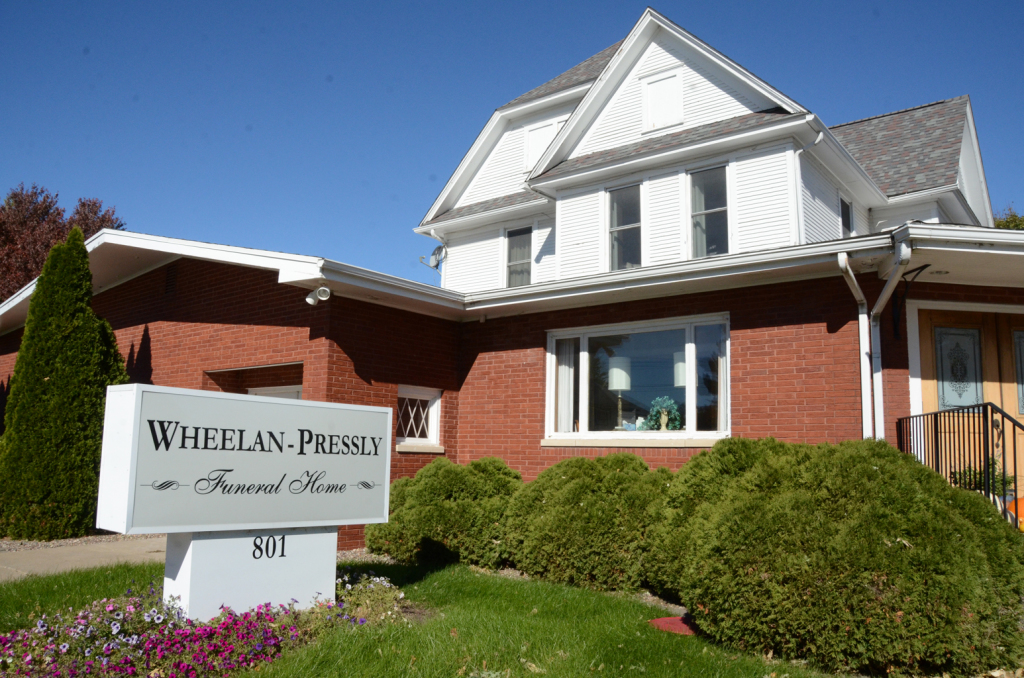 Wheelan-Pressly Funeral Home exterior: brick building with sign, green bushes, blue sky.