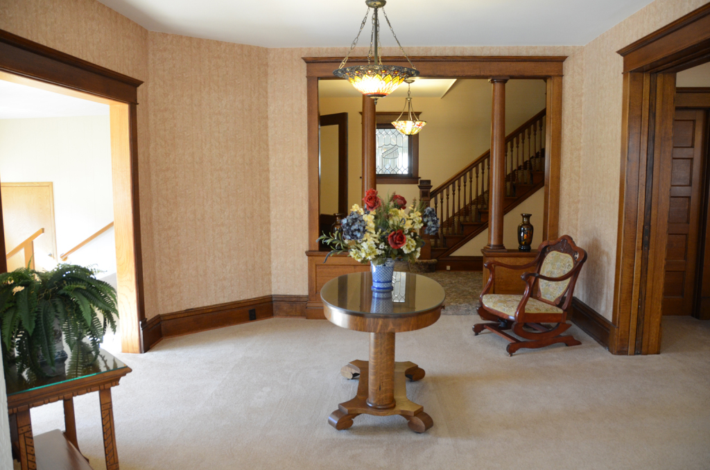 Interior of a house with a round table, rocking chair, and staircase. Light-colored walls and carpet, dark wood trim.
