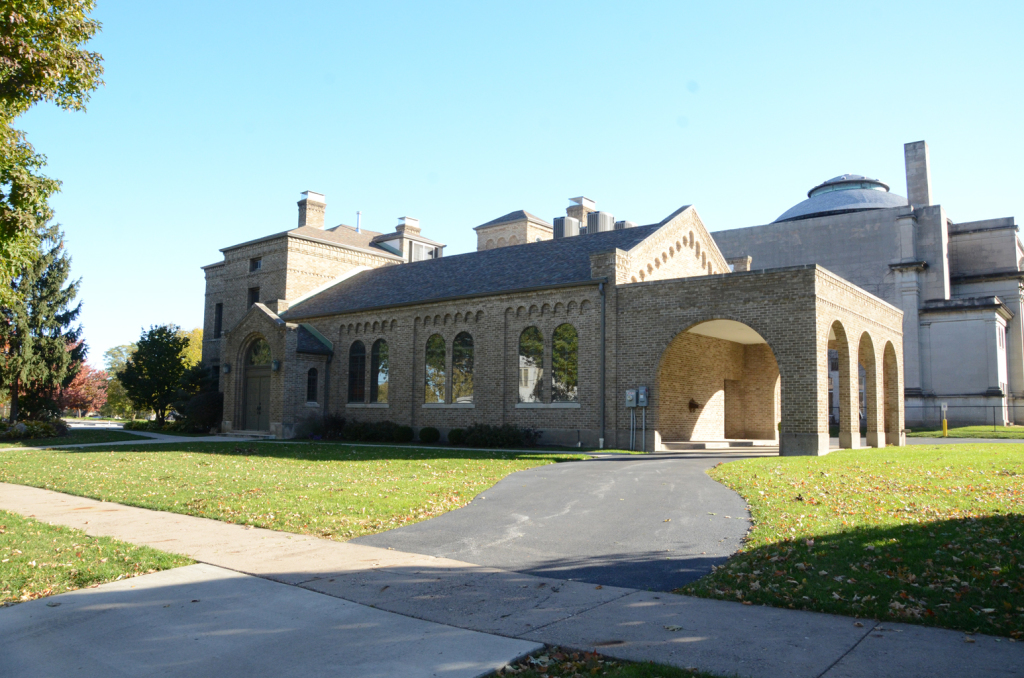 Stone building with arched entrance and a domed structure in background, on a grassy lawn with a paved path.