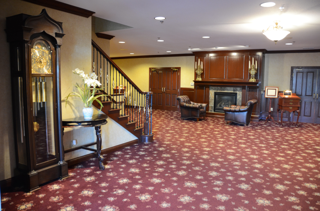 Lobby with grandfather clock, stairs, fireplace, armchairs, and patterned red carpet.