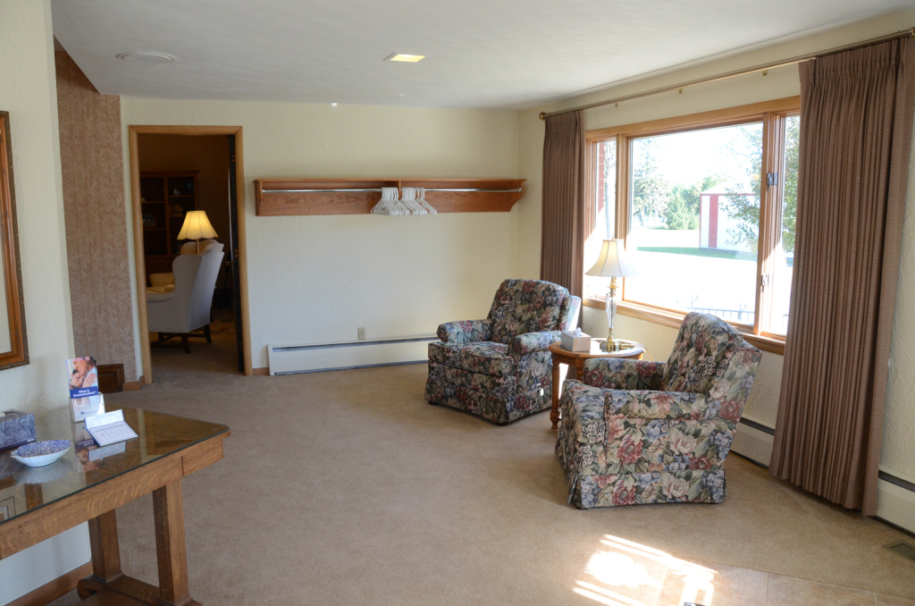Spacious living room with two patterned armchairs, large window with brown curtains, and a wooden side table.