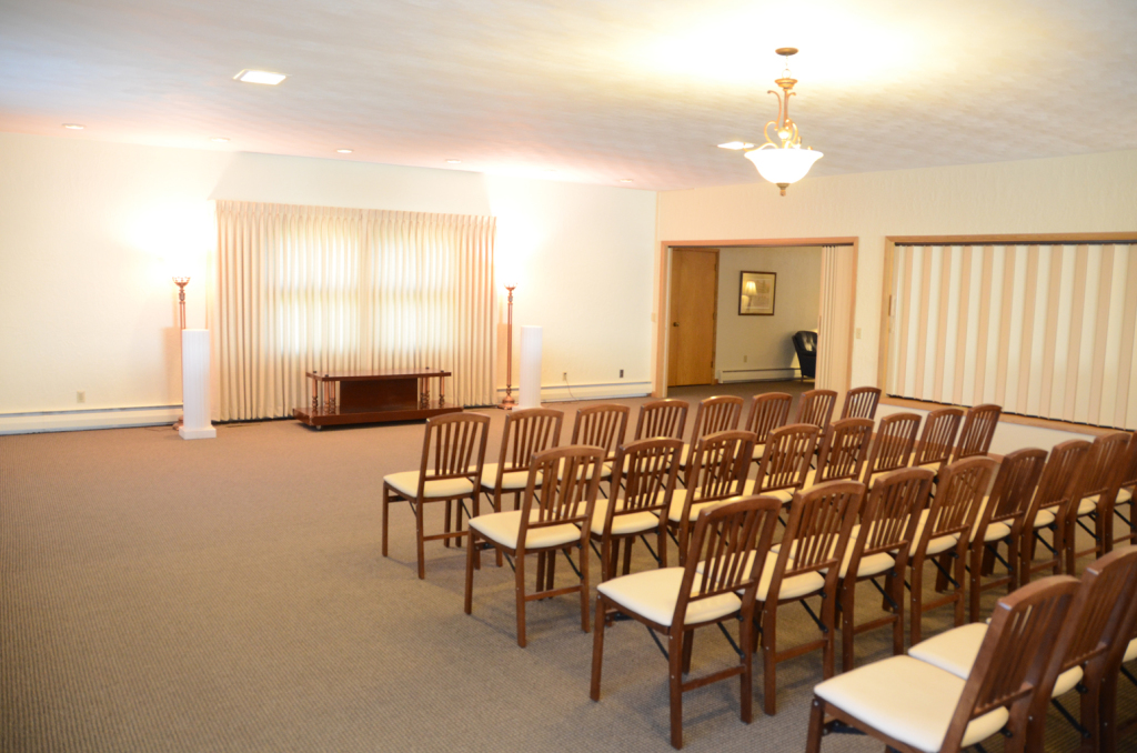 Funeral home chapel with rows of chairs facing a window and an entryway.