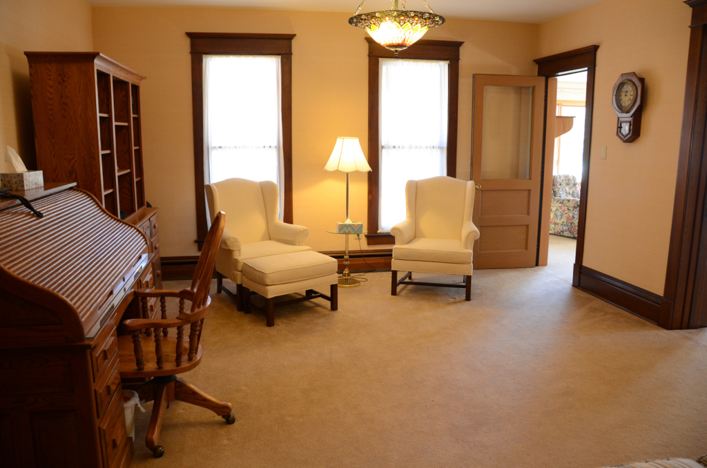Living room with desk, chairs, bookcase, windows, and doorway, light orange walls, tan carpet.