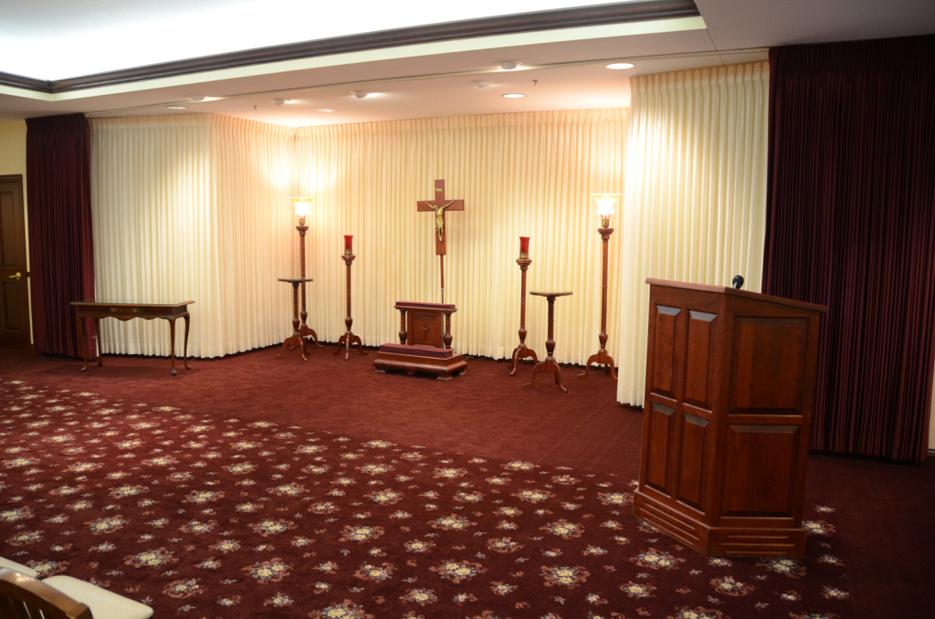 Interior of a funeral home chapel with a red carpet, altar, cross, and lectern.