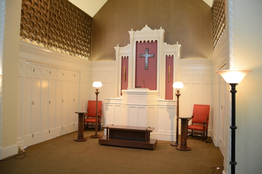 Interior view of a chapel with a cross, altar, chairs, and brown carpet.