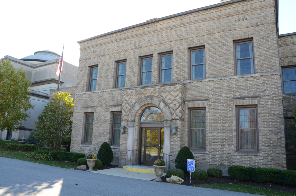 Beige brick building with arched entryway, windows, and flag. Curb appeal includes landscaping and a parking sign.