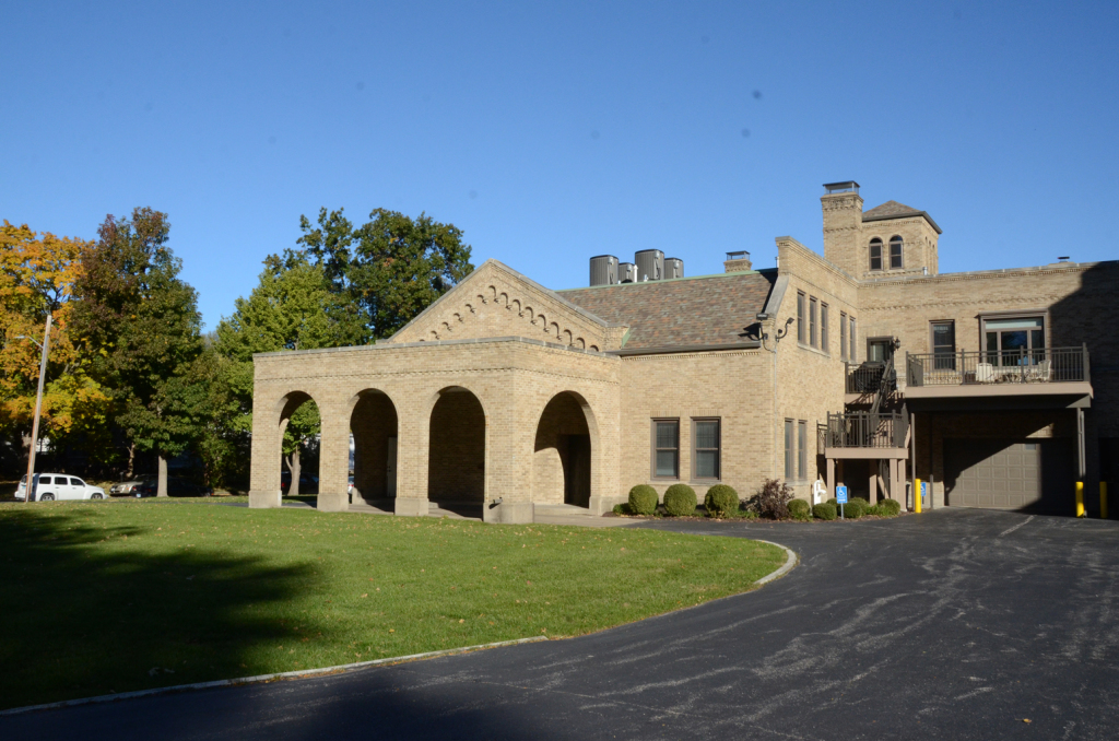 Beige brick building with arched entryway, green lawn, and winding driveway under a blue sky.