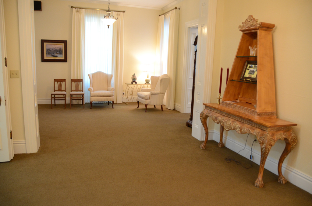 Room with light-colored walls, furniture, and curtains. A wooden cabinet stands on the right. Chairs near the window.