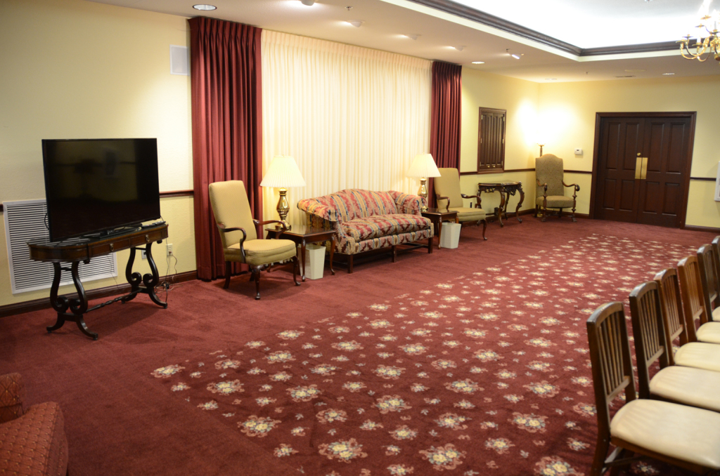 A burgundy carpeted room with a sofa, chairs, TV, and rows of chairs, lit by lamps and natural light.
