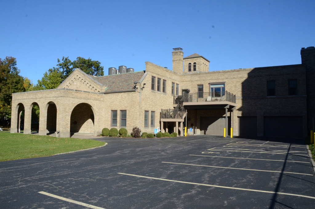 Tan stucco building with arched entryway, small tower, and parking lot. Clear blue sky.