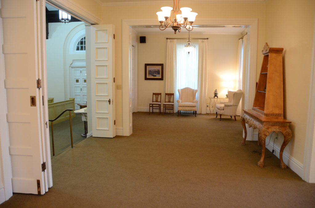 Room with tan carpet, white walls, and ornate furniture; chandelier, chairs, and open doorway to another room.