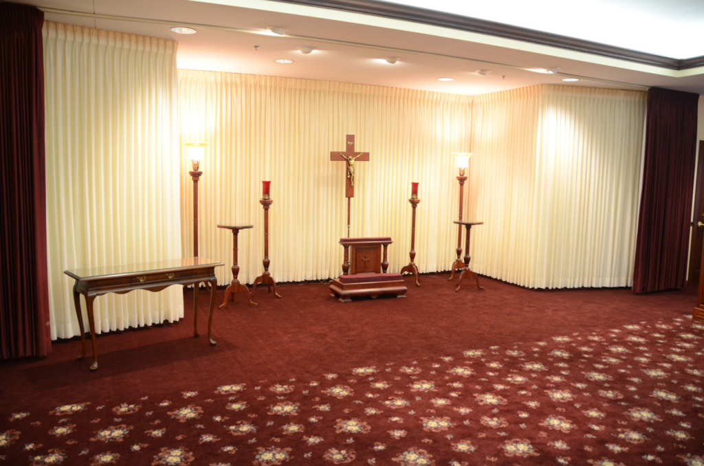 Interior of a chapel with burgundy carpet, beige curtains, a wooden cross, and candle holders.