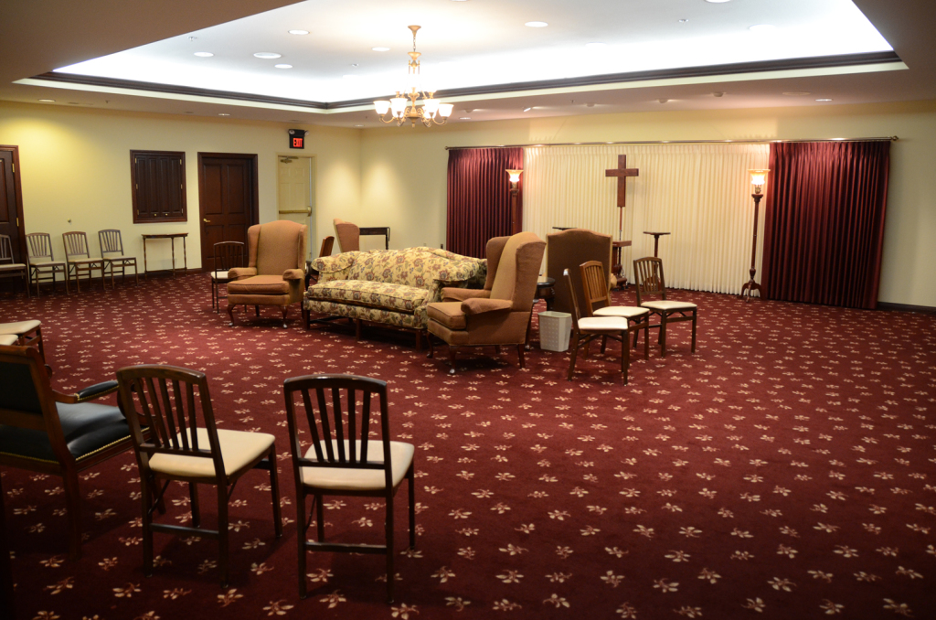 Interior of a room with chairs and a sofa arranged for a religious service; red carpet, neutral walls, and dark red curtains.
