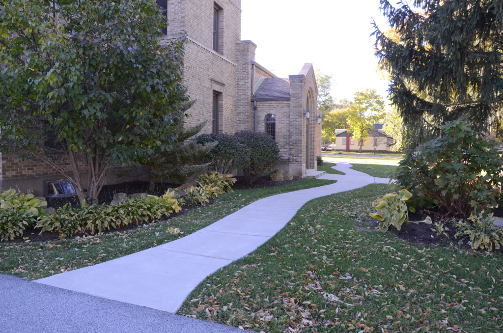 A winding concrete walkway leads past a brick building and a grassy area with trees.