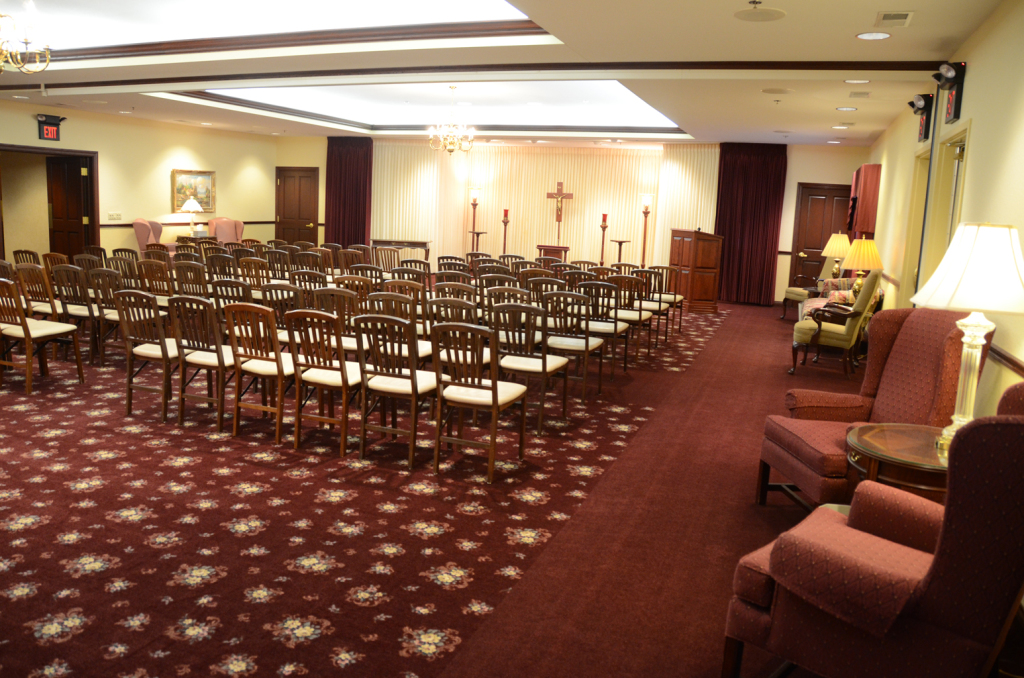 Interior view of a funeral home chapel with rows of chairs, red carpet, and a draped altar.