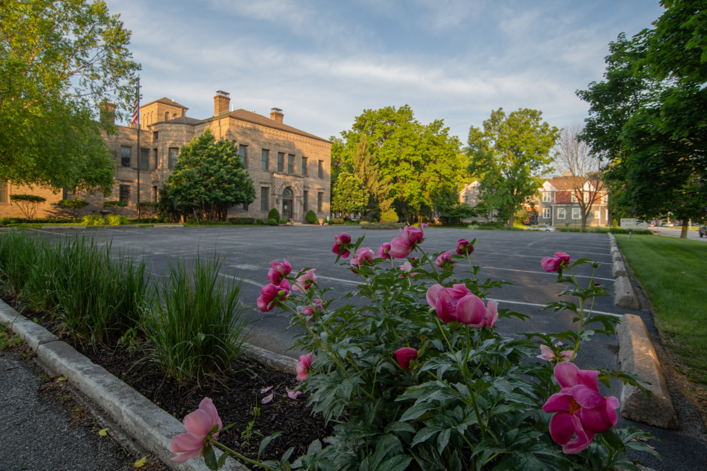 A large stone building with a parking lot and pink flowers in the foreground. Trees surround the building.