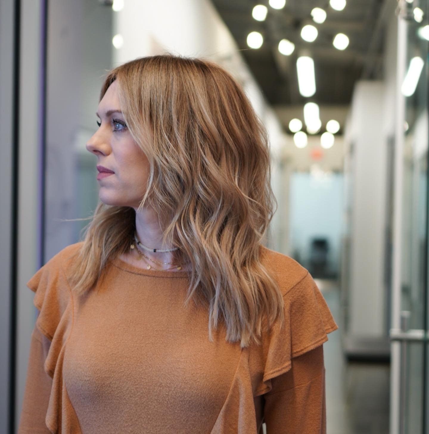 Woman in a brown sweater, looking to her left, in a hallway with bright lights.