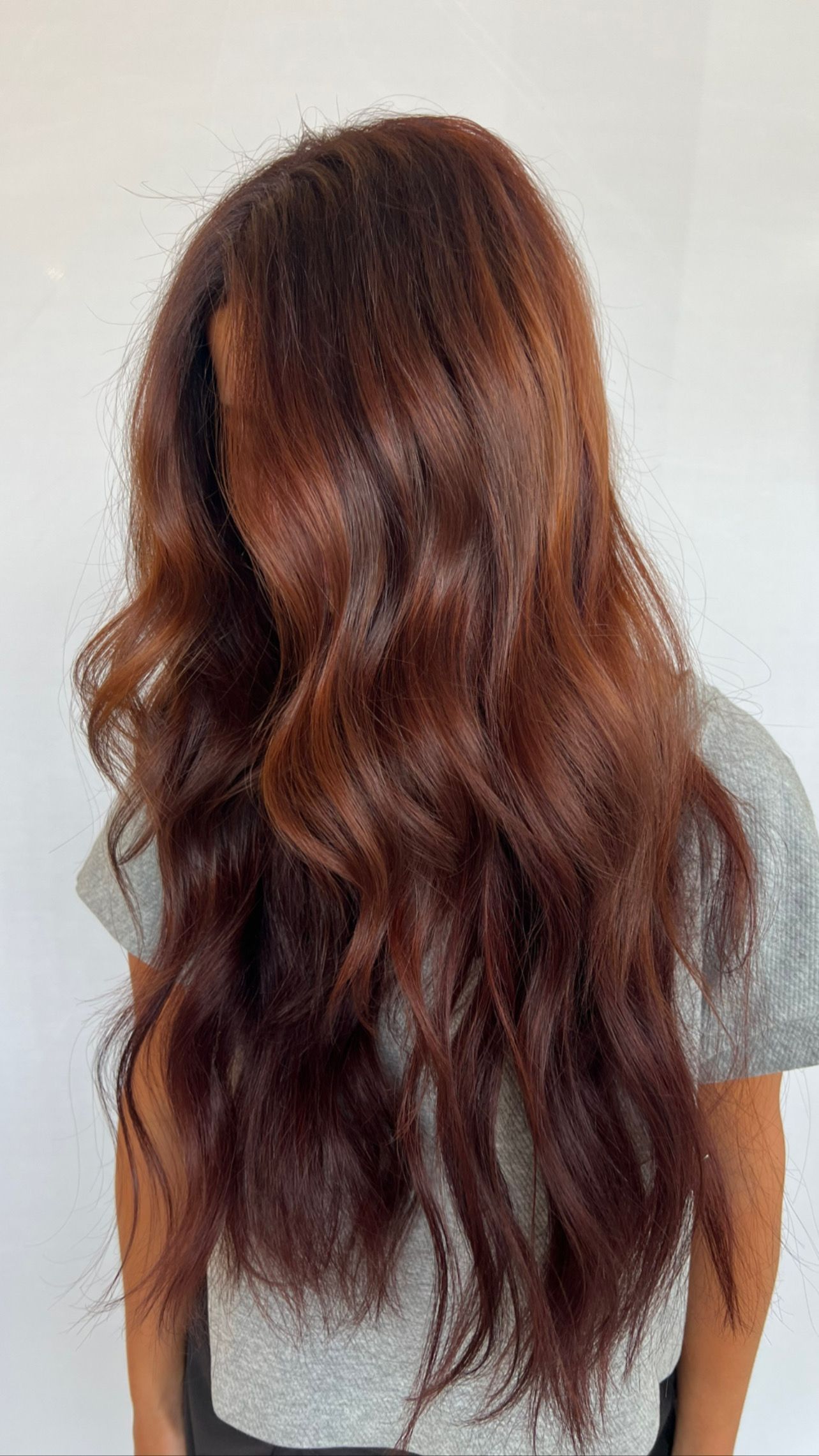 Woman with long, wavy auburn hair. Wearing a gray shirt, against a white wall.