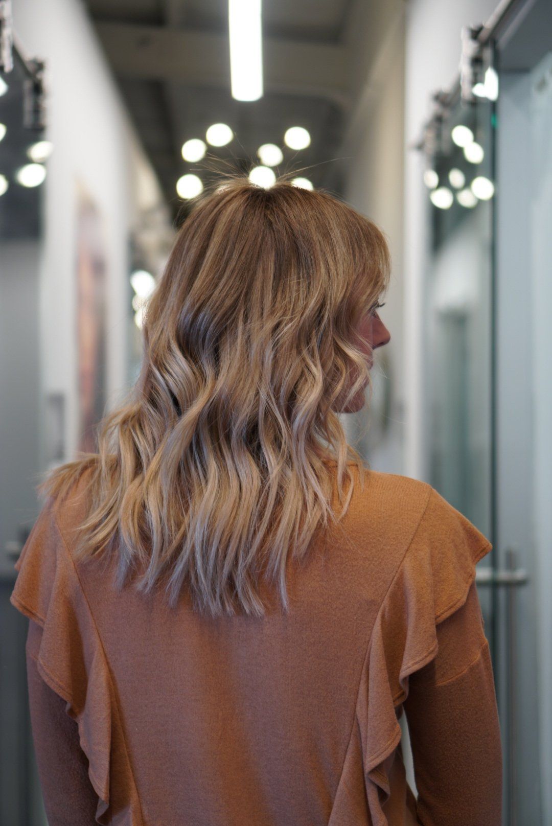Woman with blonde wavy hair, wearing a tan sweater, standing in a brightly lit hallway.